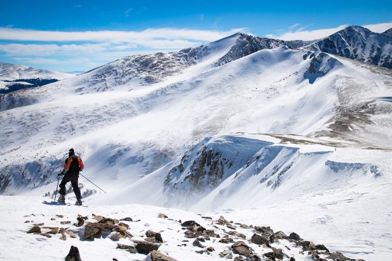 An image featuring a skier atop a mountain, poised to descend a snow-covered slope, with the bright sun illuminating the picturesque scene.